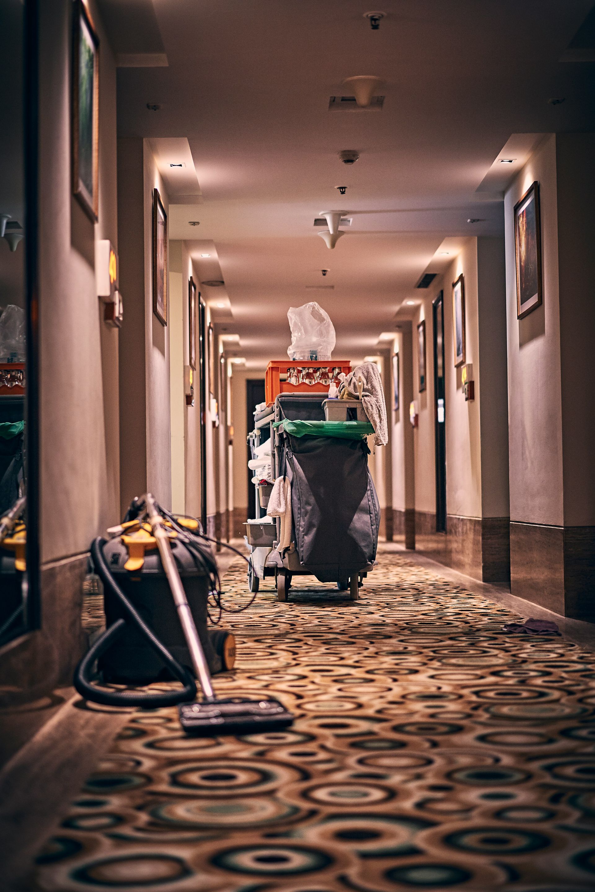 A housekeeping cart and vacuum cleaner in a long hotel corridor with patterned carpet